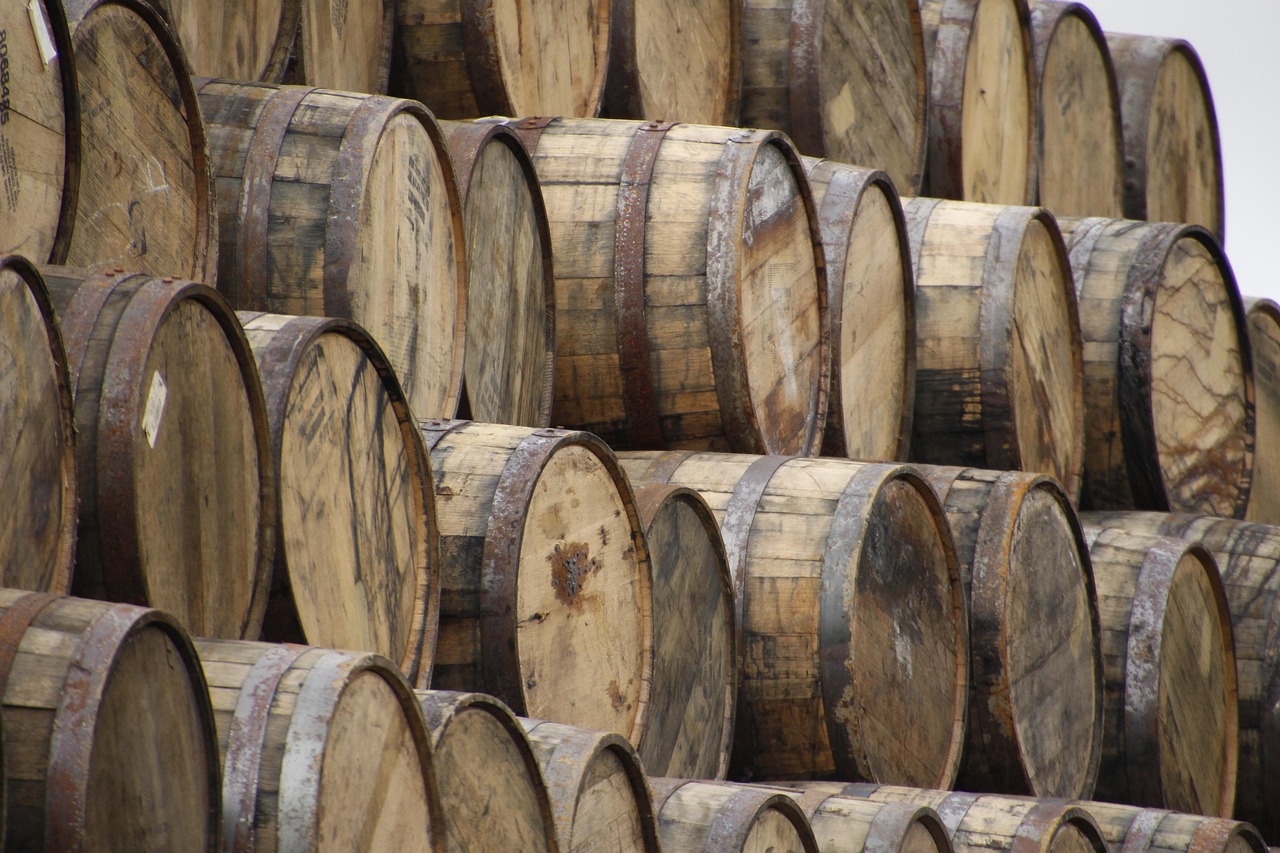 A man smiles at the camera in a cooperage with barrels and a fire behind him.