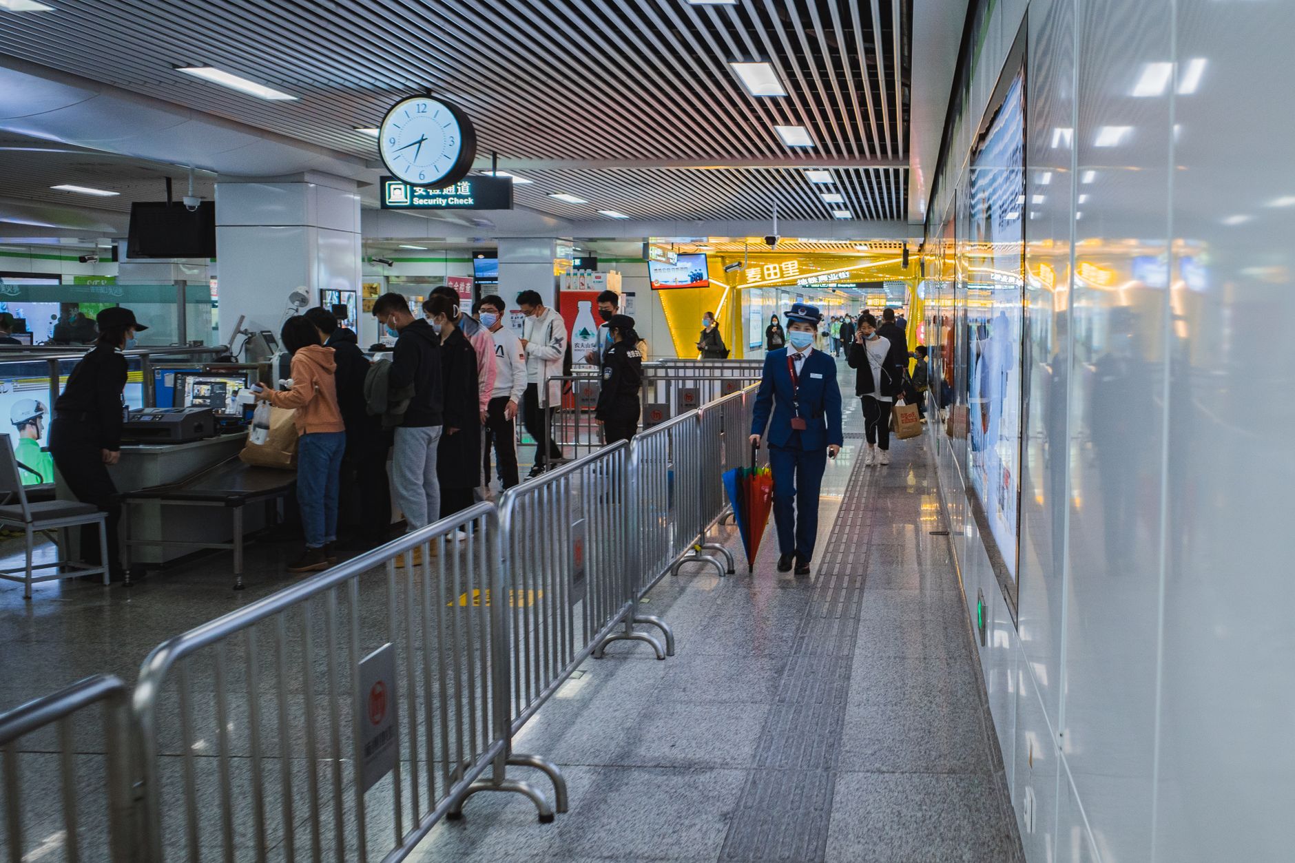 Travellers passing through an airport security queue with staff and barriers visible.
