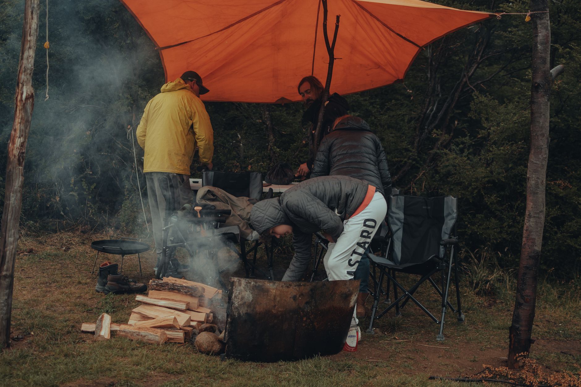 Campers in winter jackets preparing a camp kitchen beneath an orange tarp at a wet campsite.