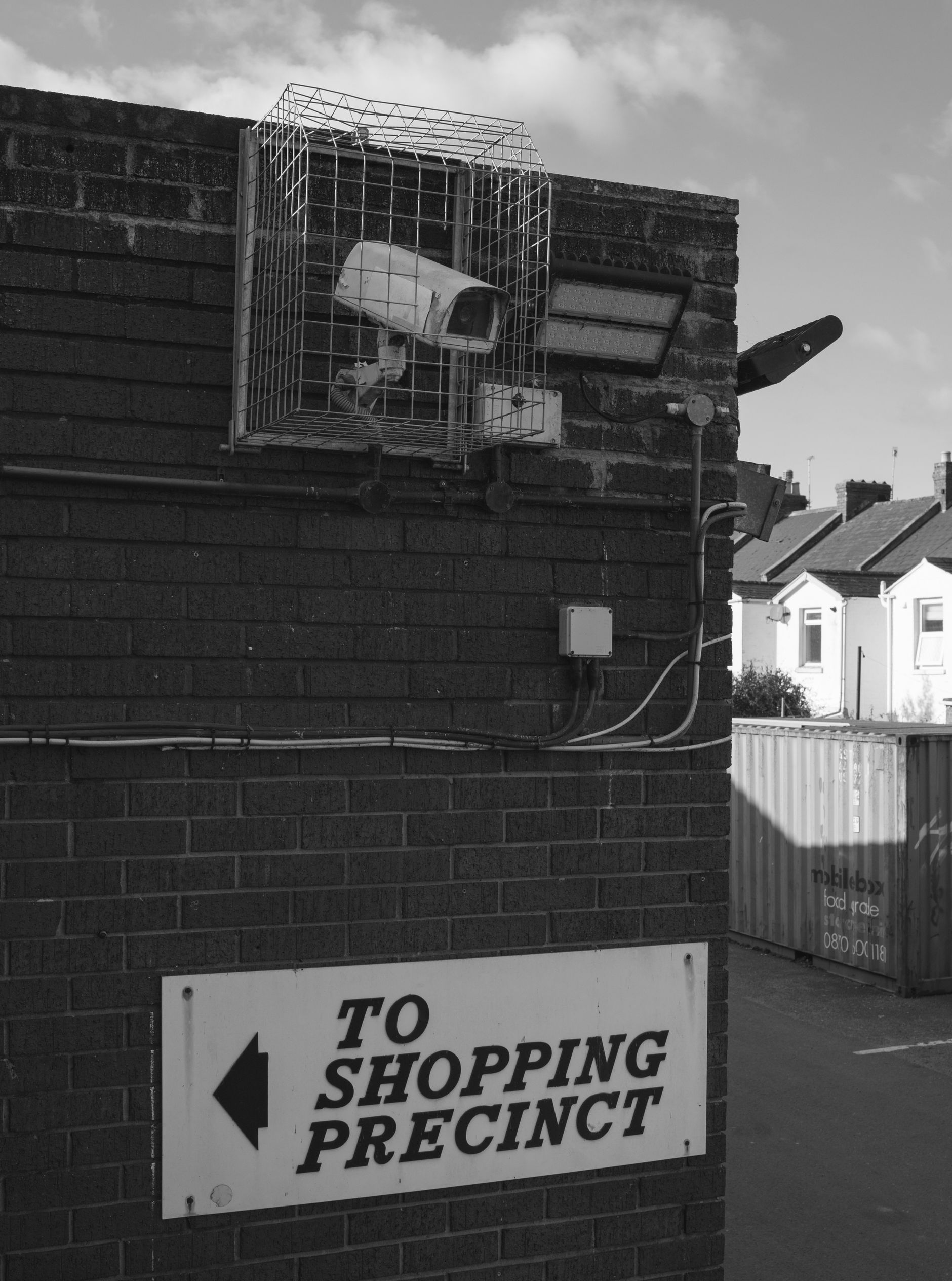 CCTV camera on a brick wall beside a shopping precinct sign.